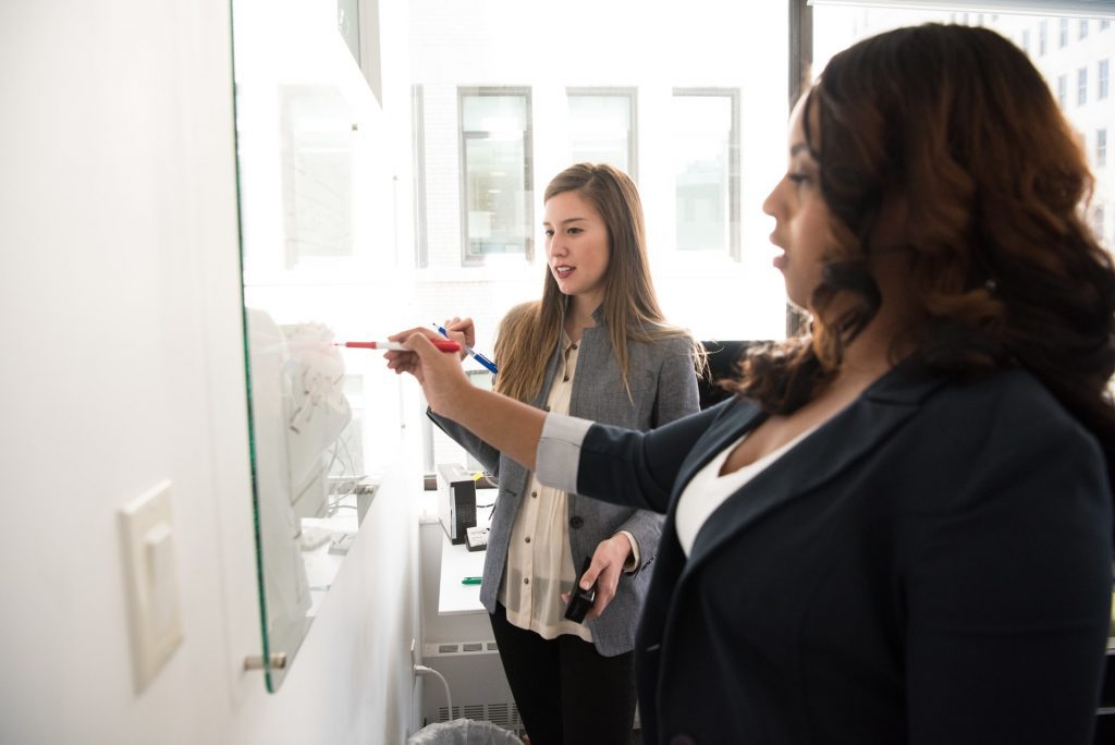 Two women are writing on a glass marker board. 