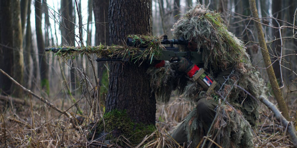 A person disguised by tree branches and grass holds a rifle in the woods