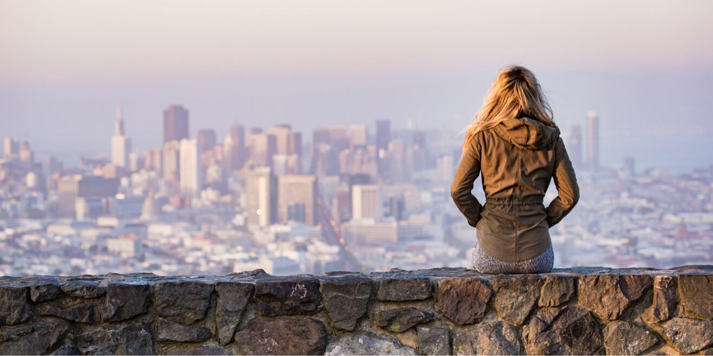 A woman sits on a stone wall and looks at a city skyline in the distance