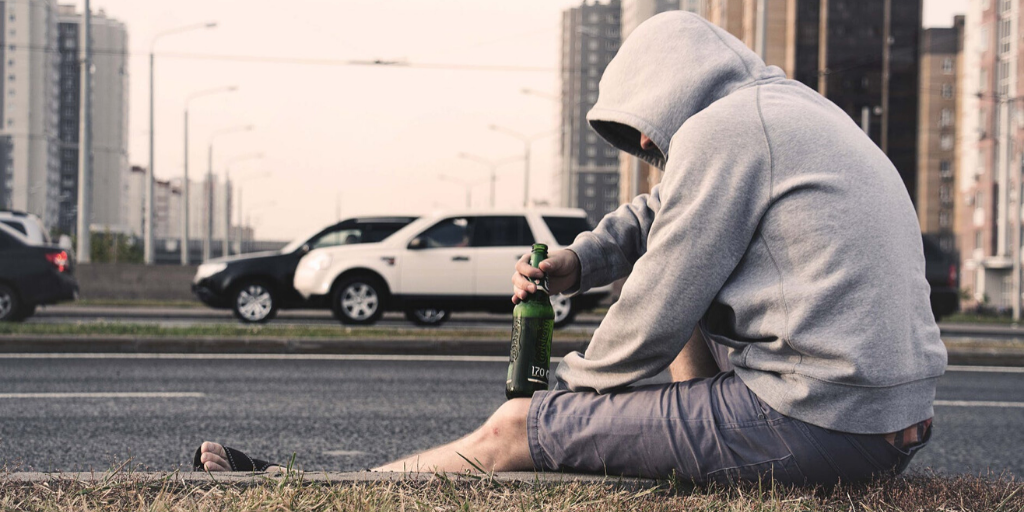 A man sits on the side of the road with a gray sweatshirt and a bottle of beer in his hand