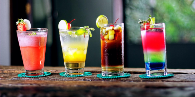 Four tropical-looking drinks with fruit garnishes are set in a line on a wooden table