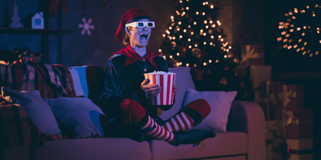A man dressed as an elf watching (perhaps) one of these four Christmas movies in English.